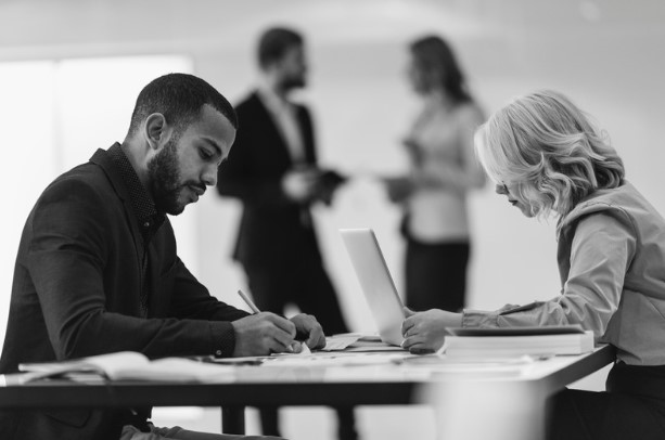 Two professionals sit at a table, working intently on documents and a laptop, while two colleagues stand in the background having a discussion. The image conveys teamwork and collaboration in a multi-agency case management environment.