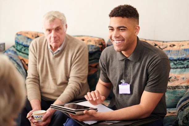 A support worker engages with an elderly man during a consultation, using a tablet to provide guidance and assistance for independent living.