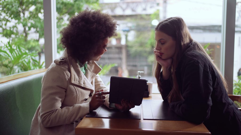 A support worker and a client are sitting together at a table in a relaxed setting, reviewing information on a tablet. The support worker, wearing a light-coloured coat, is pointing to something on the tablet while the client, dressed in dark clothing, listens attentively with her chin resting on her hand. The background shows a bright, green outdoor scene through large windows.