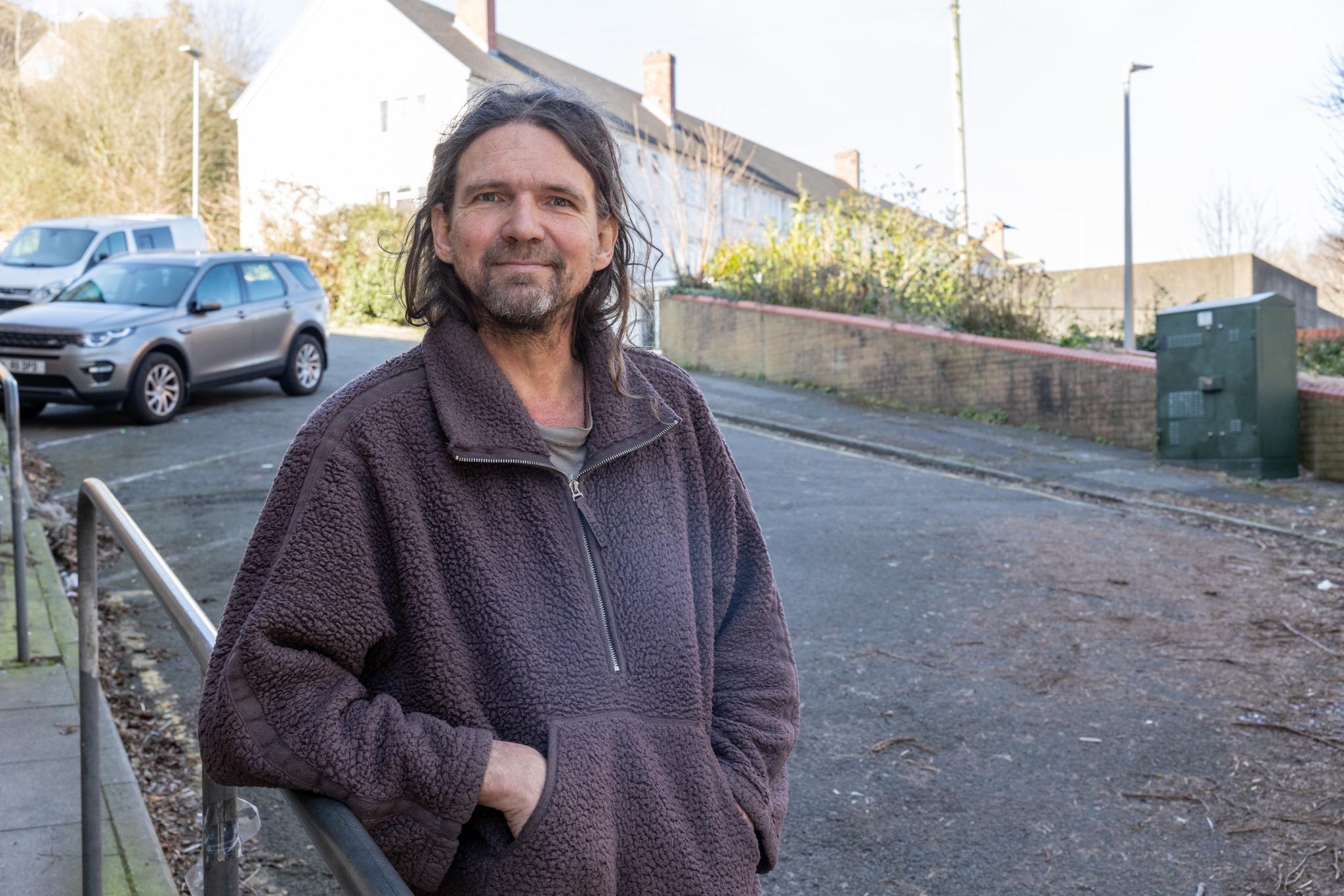 A man stands on a residential street, smiling towards the camera. The image highlights the human stories behind homelessness and housing insecurity. Photo credit: Centre for Homelessness Impact / Jeff Hubbard.
